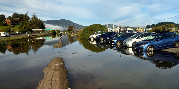 A cyclist rides through a flooded bikepath between a highway and a parking area in Sausalito, CA