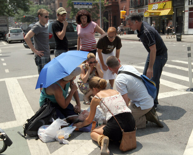 An elderly woman faints from the heat in a street intersection on the Upper East Side. A group of concerned bystanders is gathered around her attending to her needs, including someone holding a blue umbrella to shade her.
